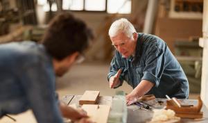 Dans un atelier d'ébénistes, un homme âgé montre une tâche à un jeune homme flouté