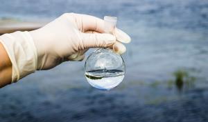 Une main gantée tient un contenant empli d'eau avec en fond un étendue d'eau