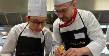 Un cuisinier montre un geste pro à une apprentie