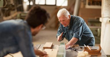 Dans un atelier d'ébénistes, un homme âgé montre une tâche à un jeune homme flouté