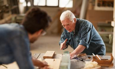 Dans un atelier d'ébénistes, un homme âgé montre une tâche à un jeune homme flouté