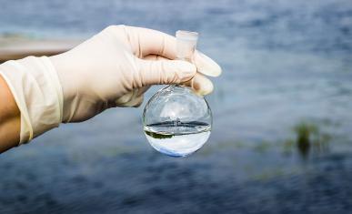 Une main gantée tient un contenant empli d'eau avec en fond un étendue d'eau