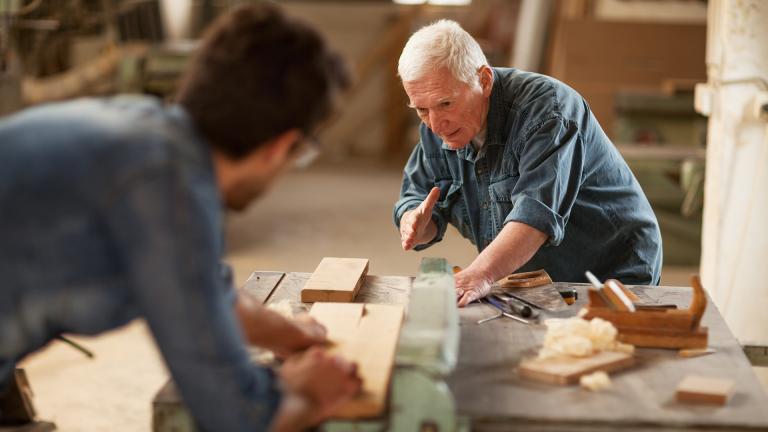 Dans un atelier d'ébénistes, un homme âgé montre une tâche à un jeune homme flouté