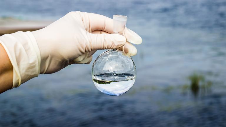 Une main gantée tient un contenant empli d'eau avec en fond un étendue d'eau